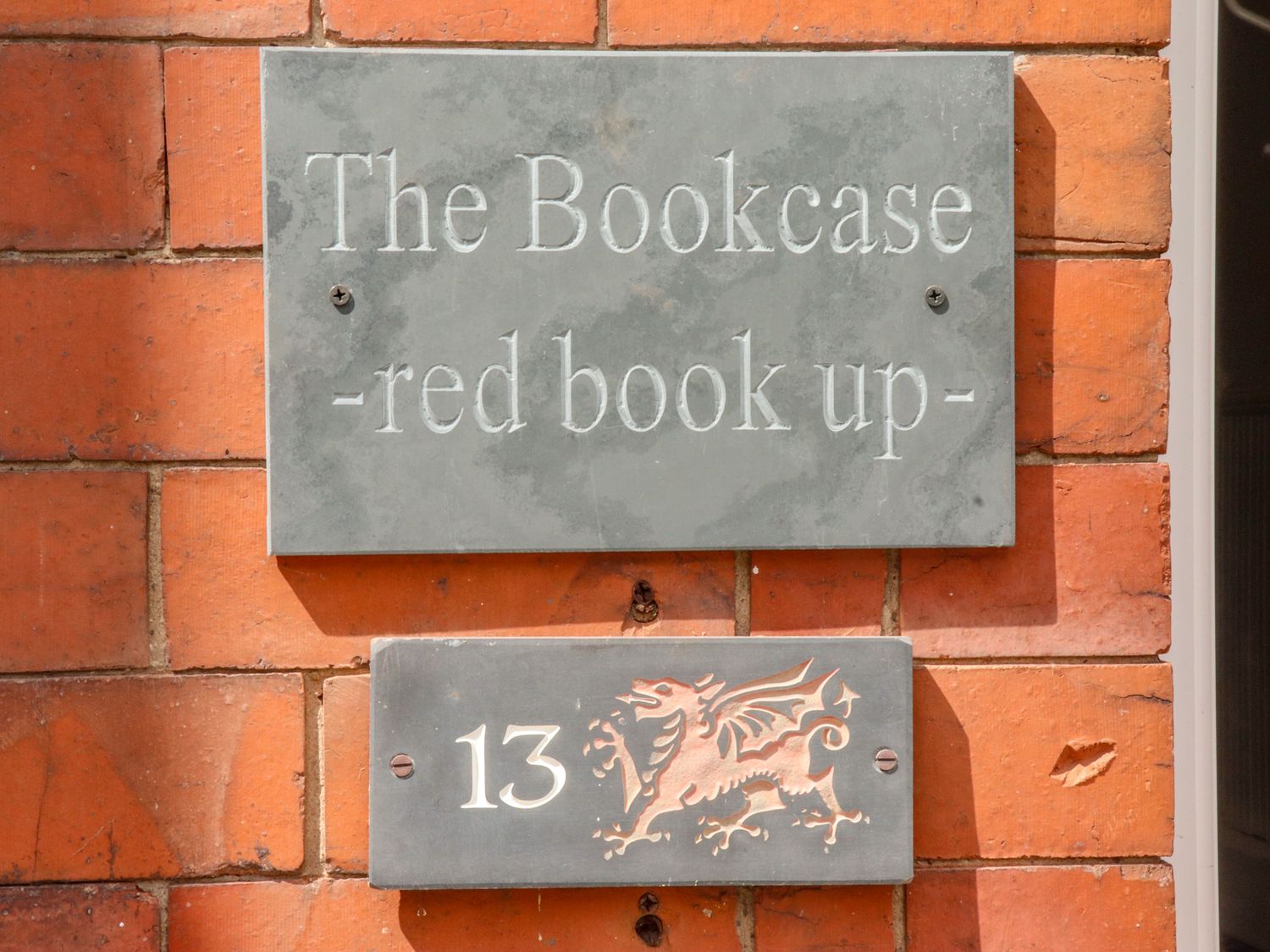 A brick wall with two gray plaques one with text the bookcase red book up and another with number 13 and a dragon emblem at The Bookcase in Colwyn Bay