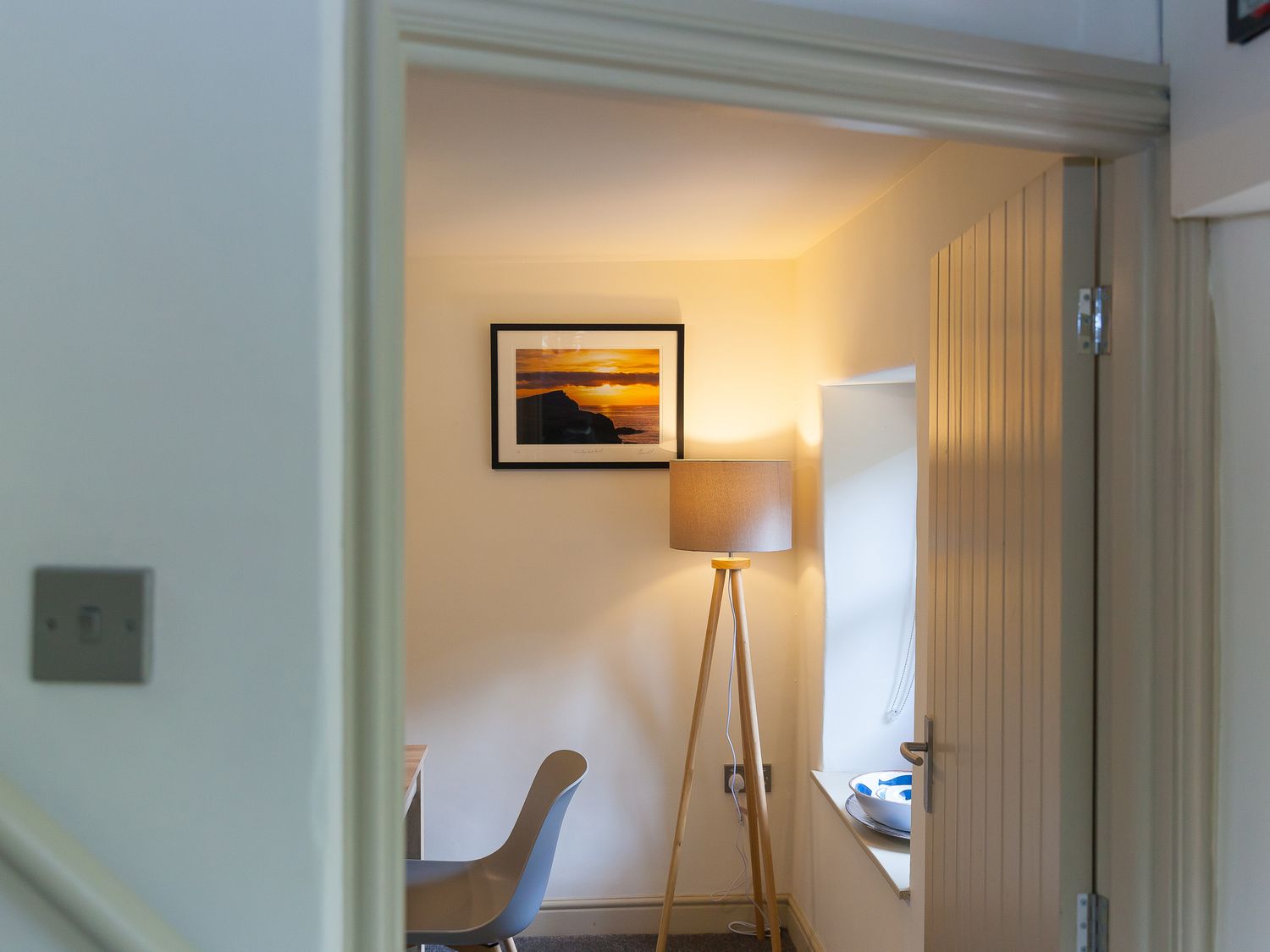 A room with a grey chair a wooden tripod floor lamp a framed sunset picture and a window with a bowl on the sill at Trewolla Cottage in Newquay