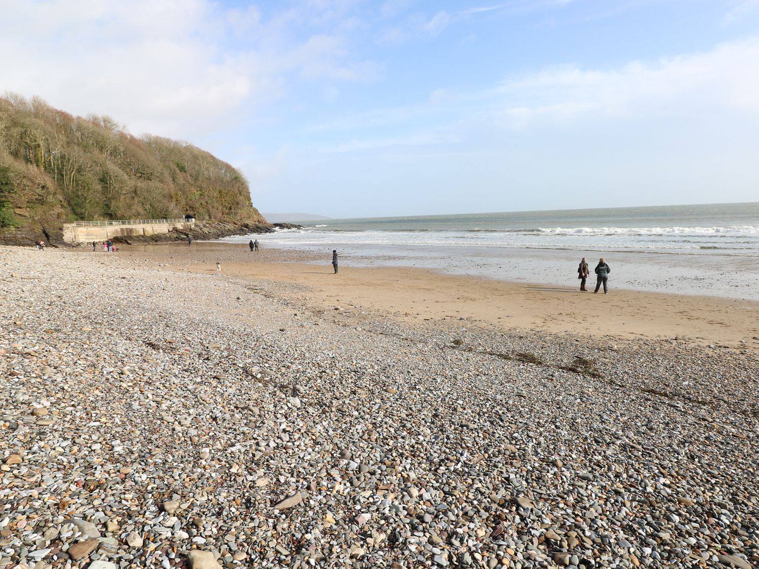 A pebble and sandy beach with people walking near the shoreline and a wooded hill in the background at Pinewood in Saundersfoot