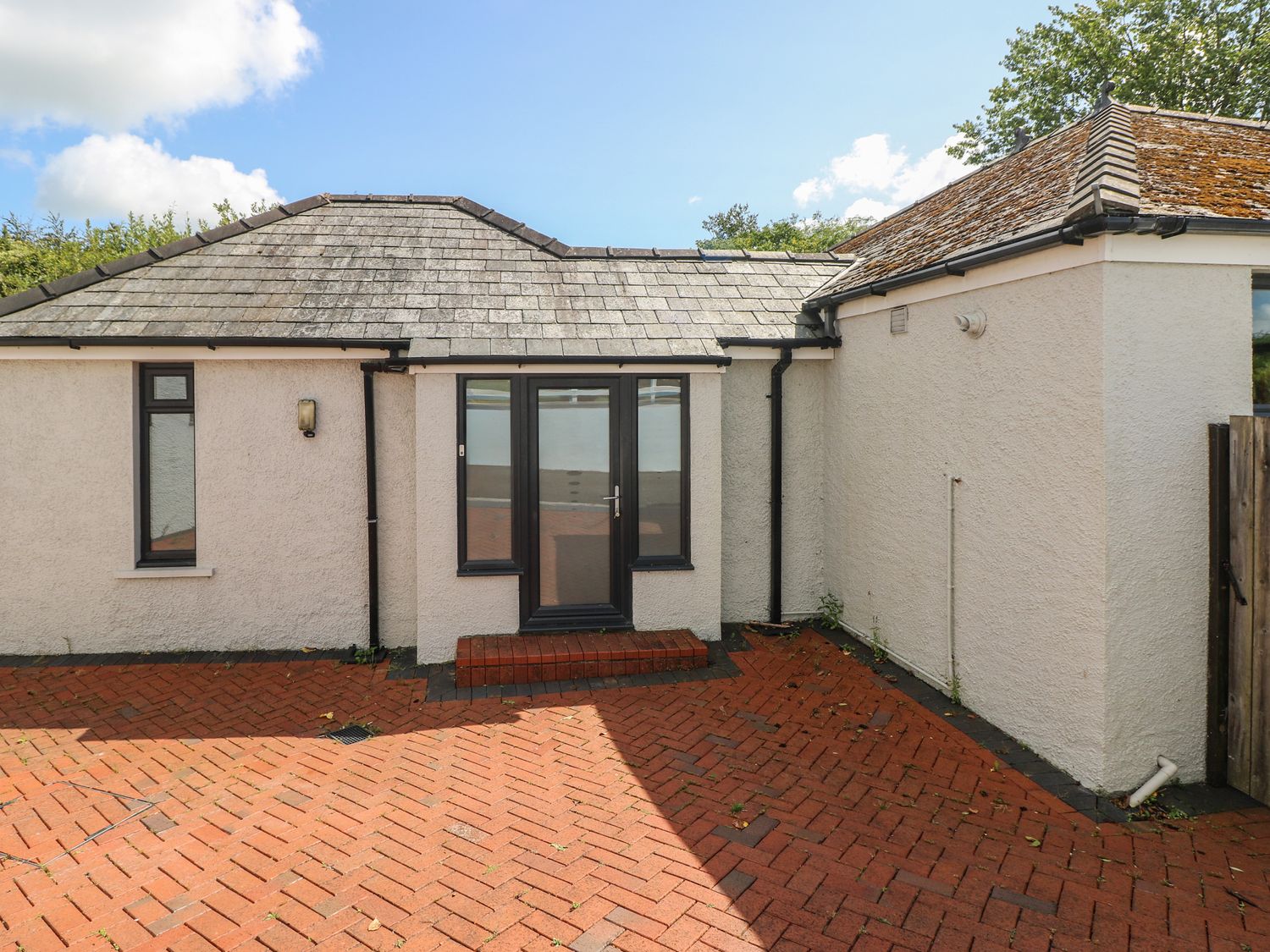 A rear exterior view of a single-story house with a tiled roof and black framed glass door opening onto a red brick patio at Pinewood in Saundersfoot