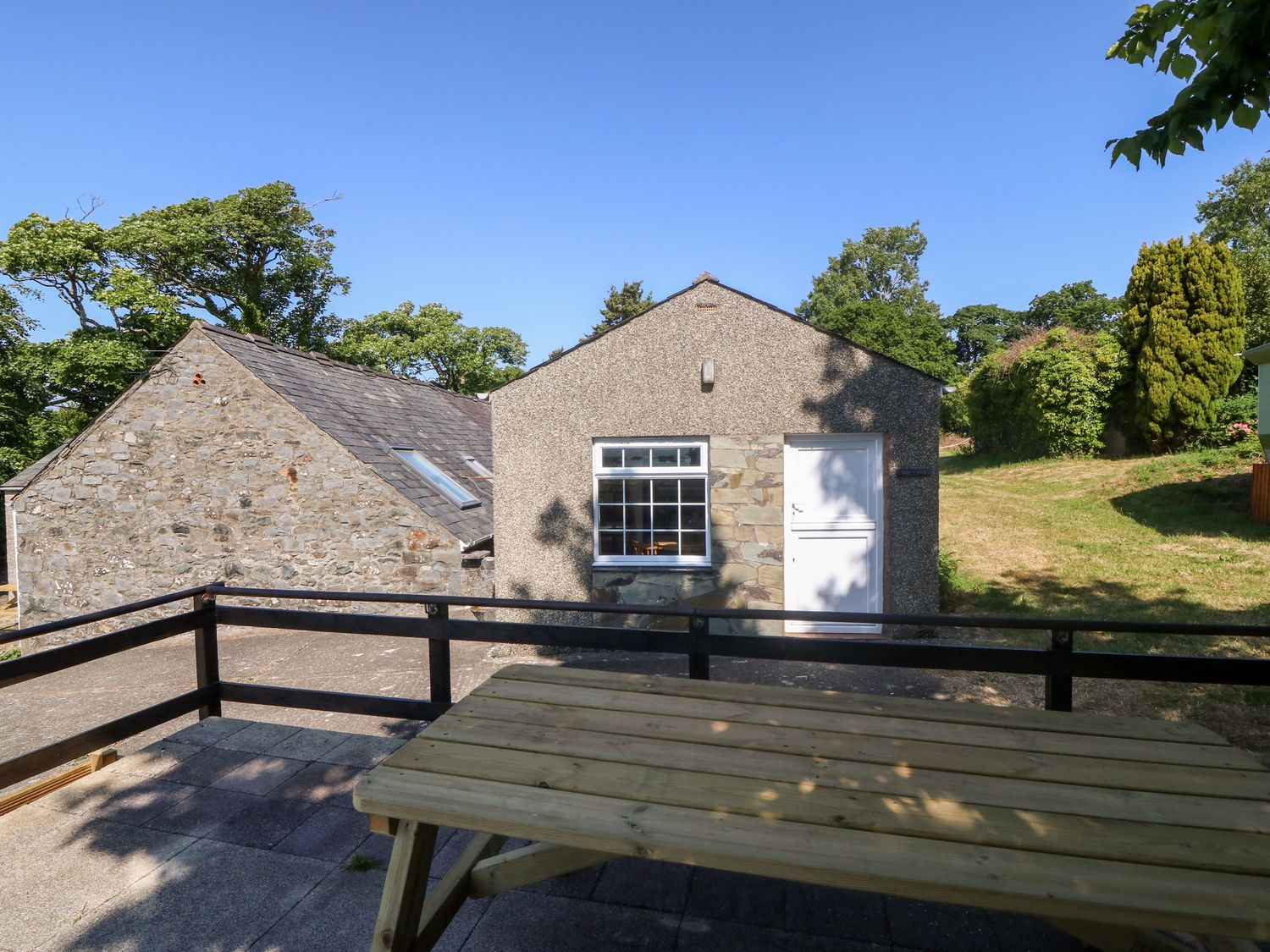 An outdoor patio area with a wooden picnic table and railing next to a building with stone and stucco walls at Min Y Mor in Llanystumdwy near Criccieth