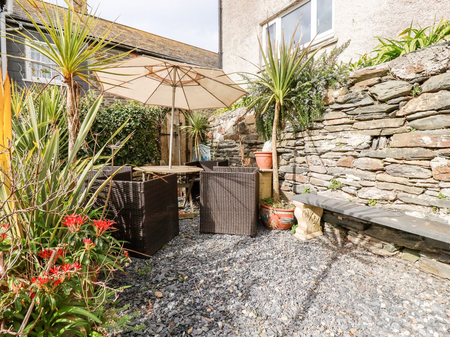 An outdoor patio area with a stone wall a table with an umbrella four chairs and various potted plants at The Moonbeach Cabin in Gorran Haven