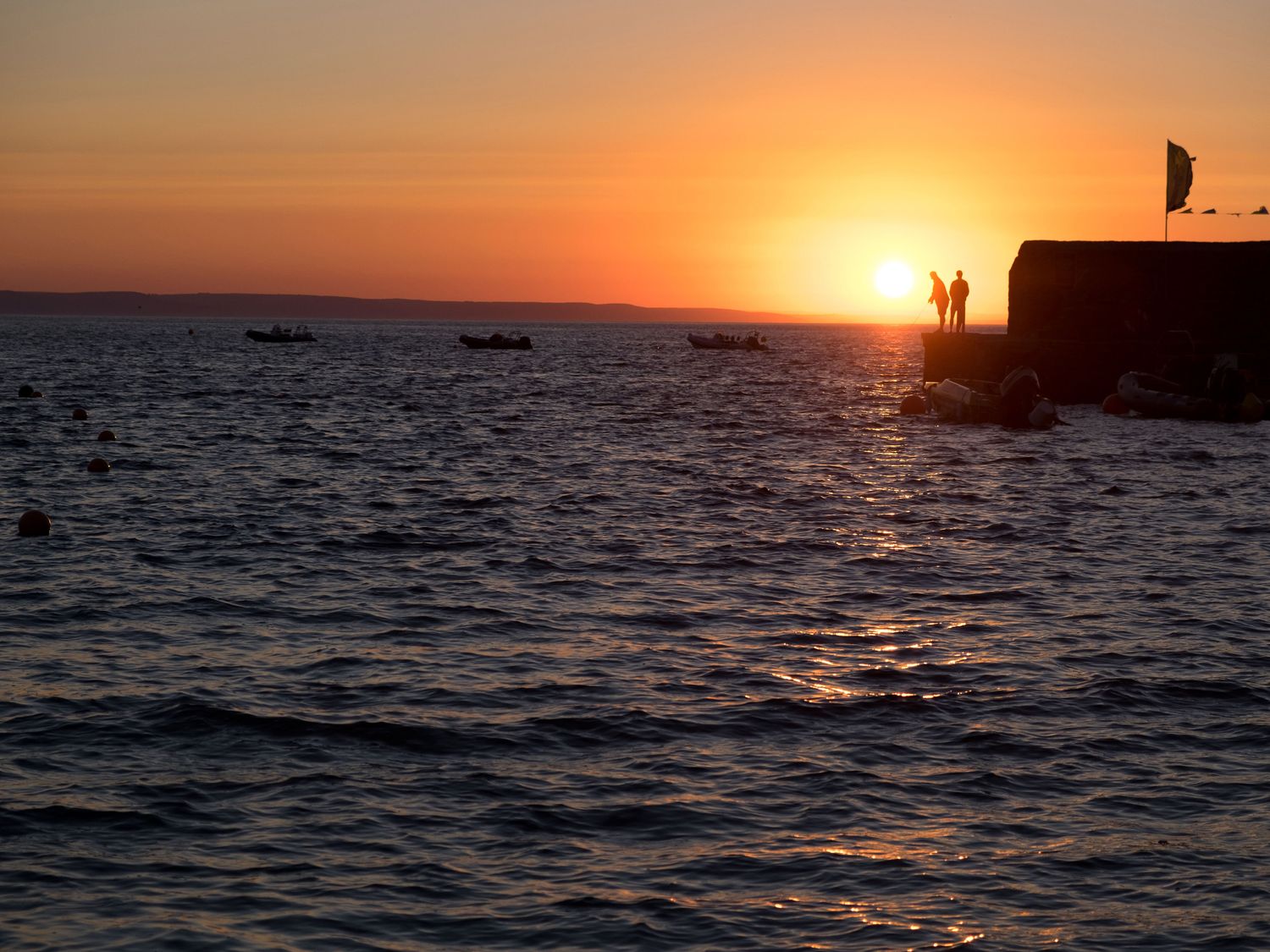 A sunset over the sea with boats and two people standing on a pier at The Moonbeach Cabin in Gorran Haven