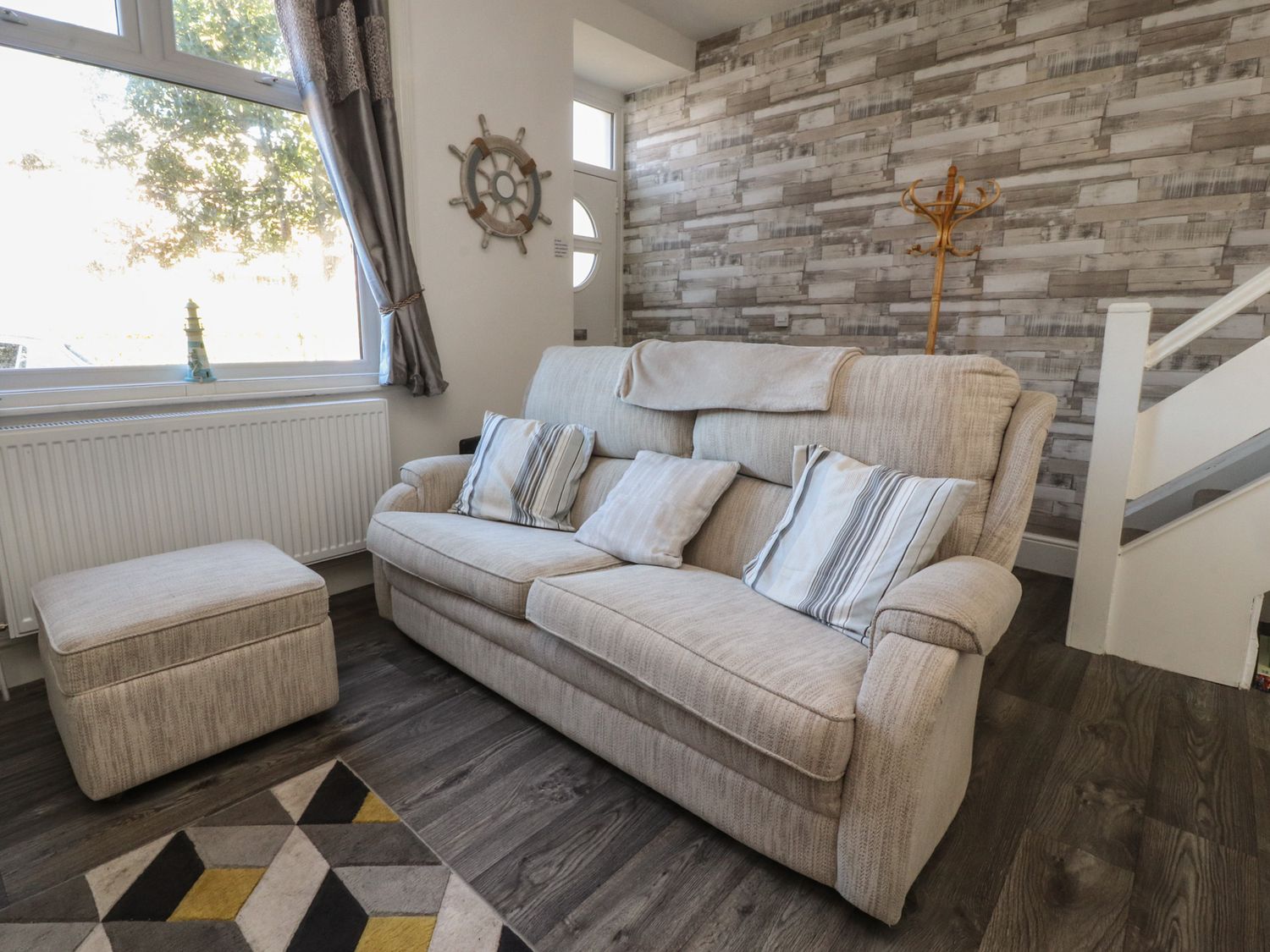 A living room with a beige sofa with cushions an ottoman a patterned rug a wooden coat rack and a window with curtains at 23 Dock Road in Lytham St Annes