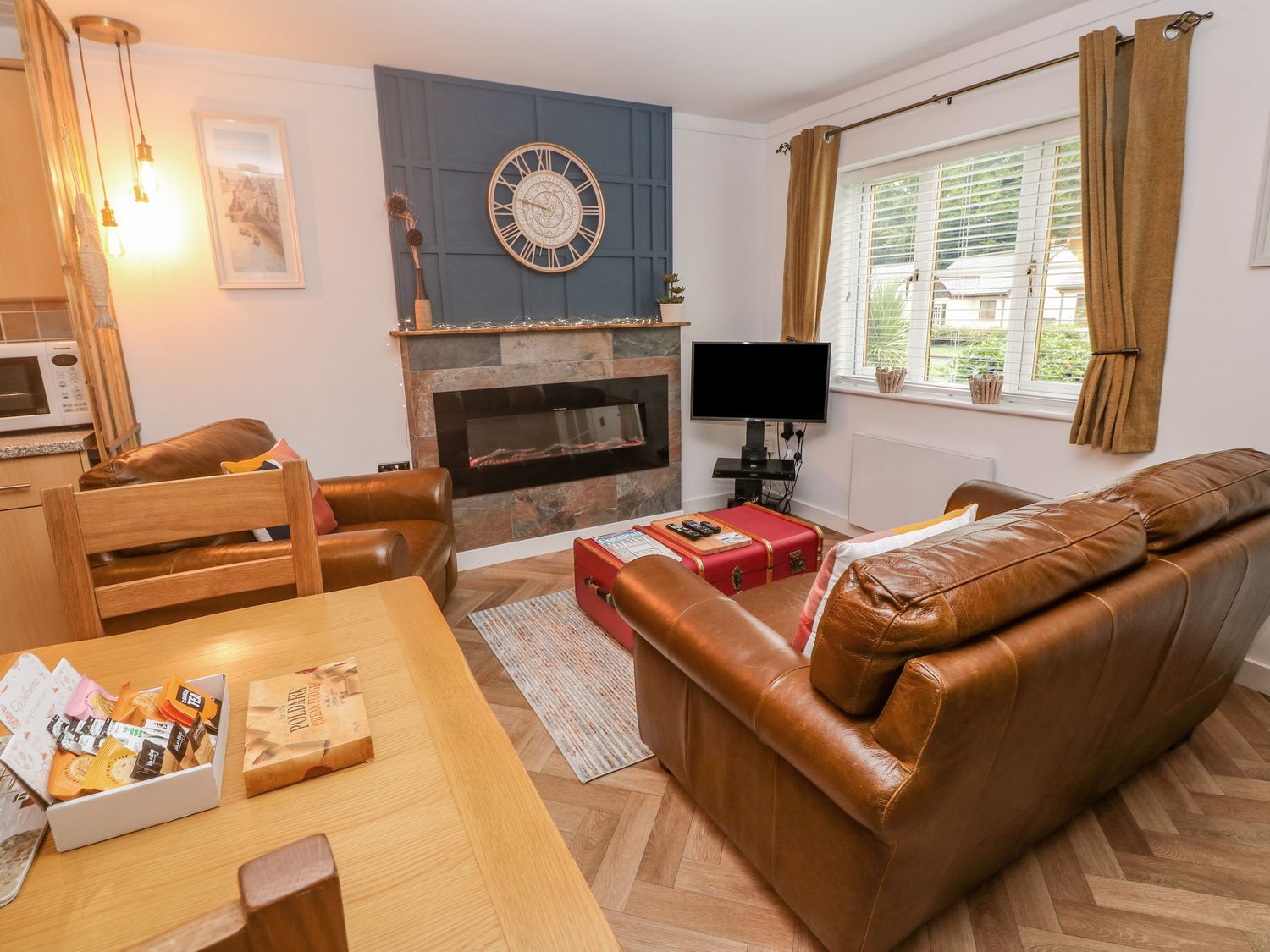 A living room with brown leather sofas a fireplace with a large clock above a TV on a stand near a window with curtains at Number 17 Bell Cottage in Camelford