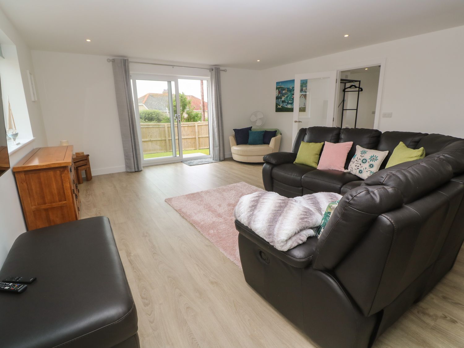A living room with a black leather sofa, a wooden cabinet, a round beige chair, and sliding glass doors at 3 Praa Cove in Praa Sands