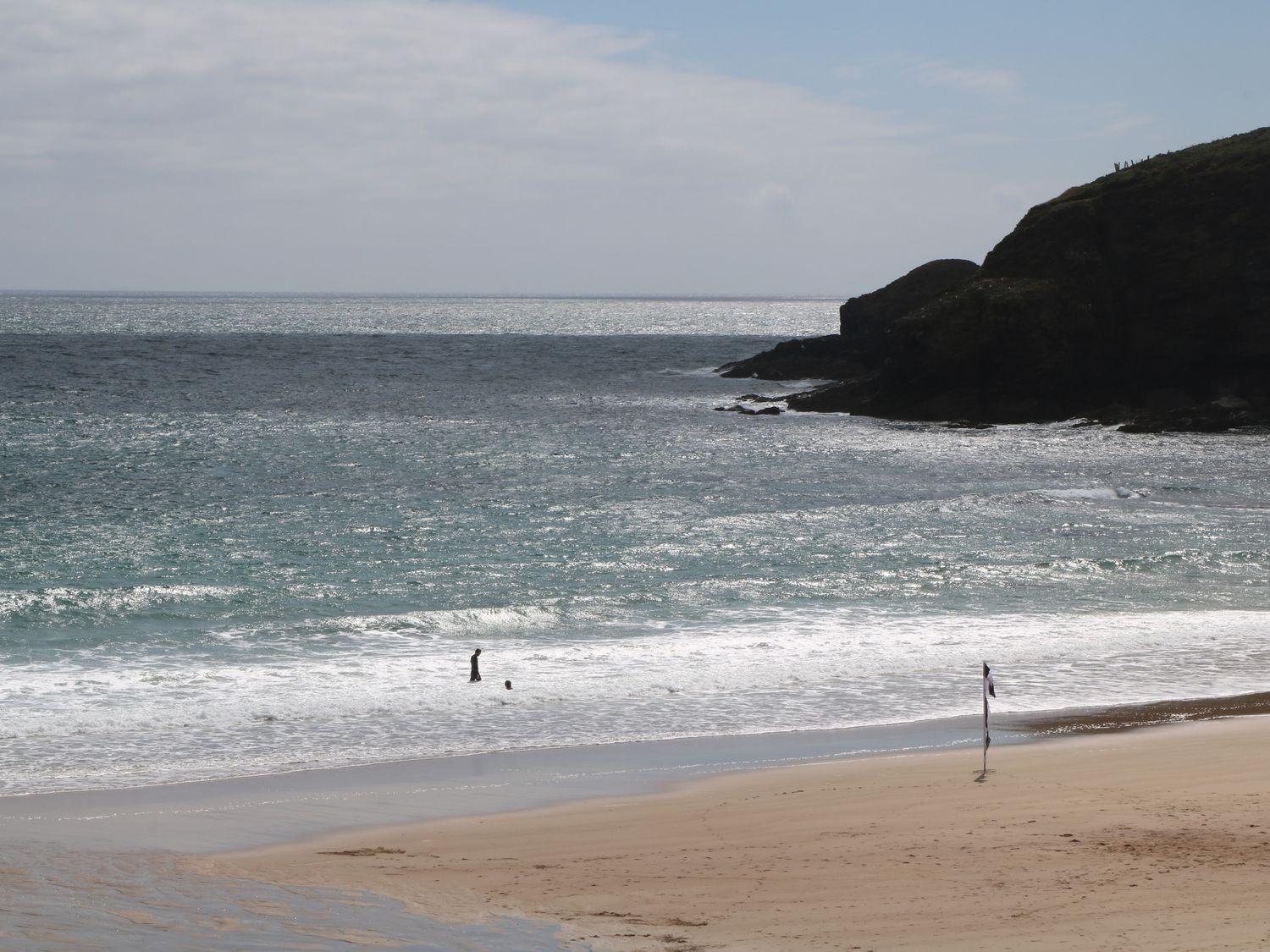 A beach with two people in the water and a flag on the sand near rocky cliffs at 3 Praa Cove in Praa Sands