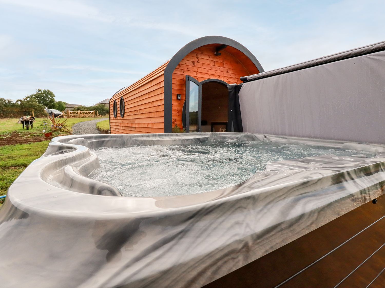 An outdoor hot tub with bubbling water next to a wooden pod with circular windows and an open door at Stargaze in Ponsanooth