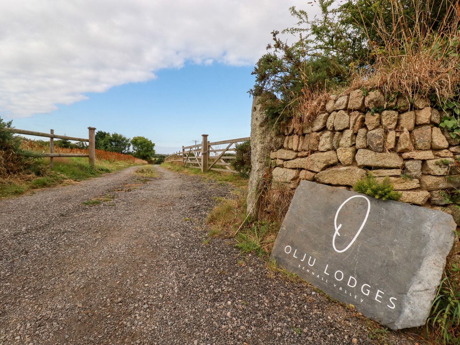 A gravel road with wooden fences on the sides and a stone wall with a sign reading Olju Lodges Kennall Valley