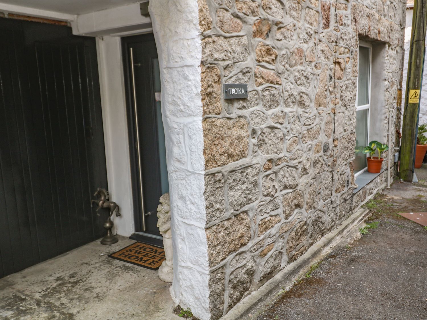 The exterior stone wall of a building with a dark door a welcome mat horse sculpture and potted plants at Troika in Newlyn