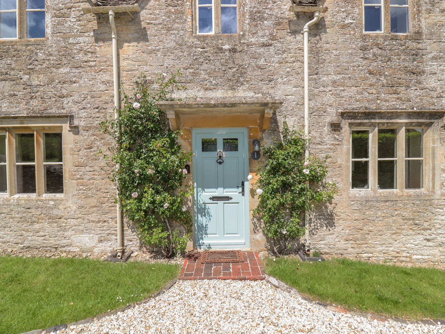 A stone house exterior with a light blue front door flanked by climbing plants and windows on either side at No 1 Eastington Eastington near Northleach