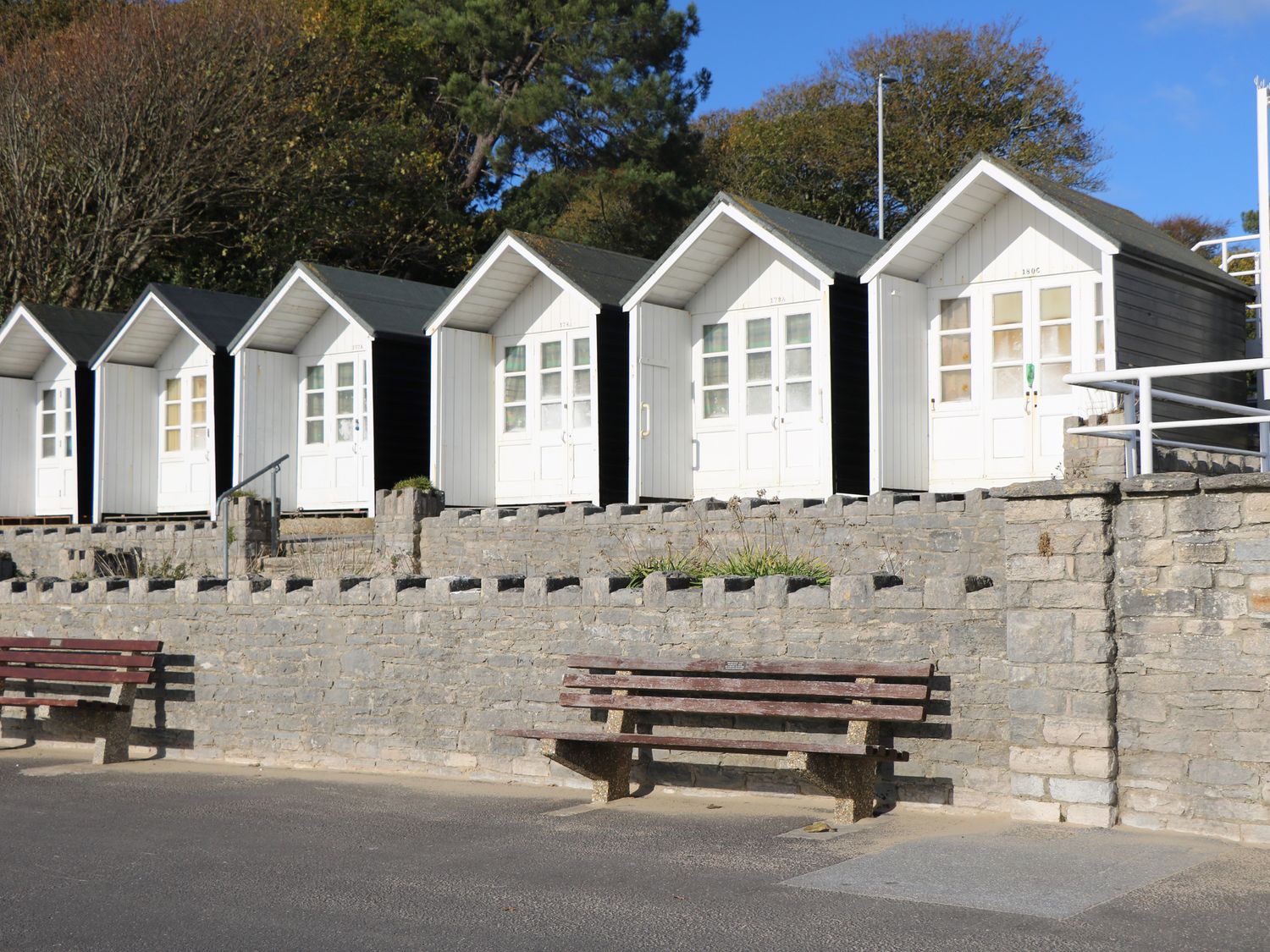 A row of small white and black beach huts behind a stone wall with wooden benches in front at 1 Charlotte Close Talbot Village Poole