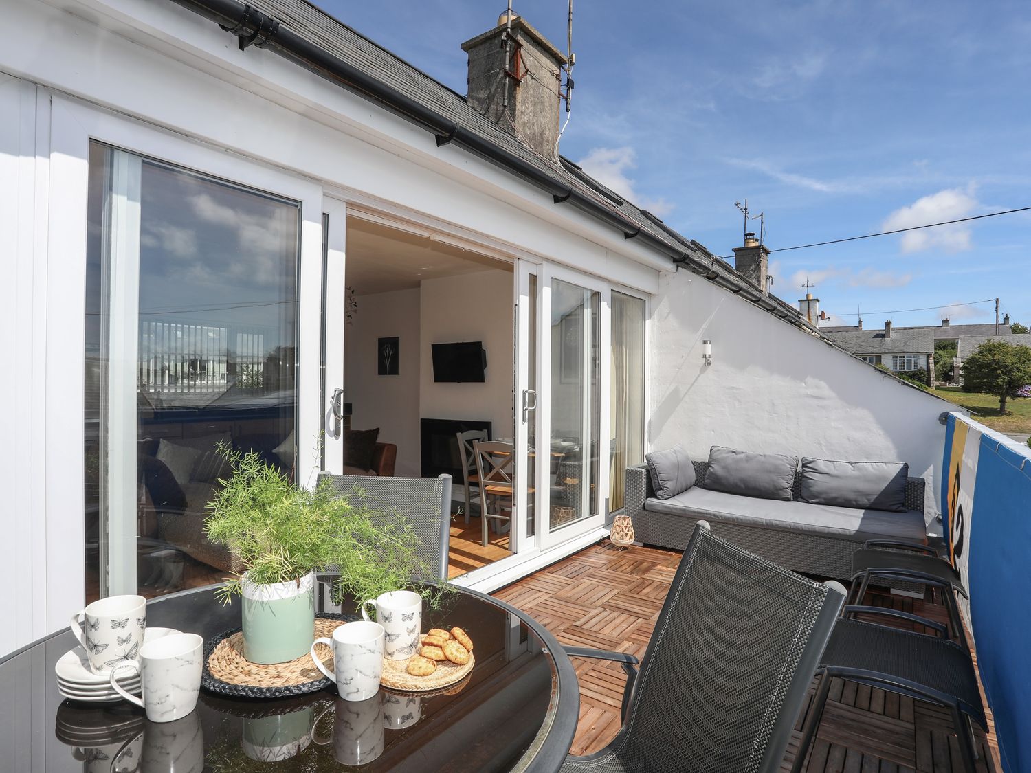 A balcony with a round table set with mugs and a plant and a sofa with cushions at 21 Cae Du in Abersoch