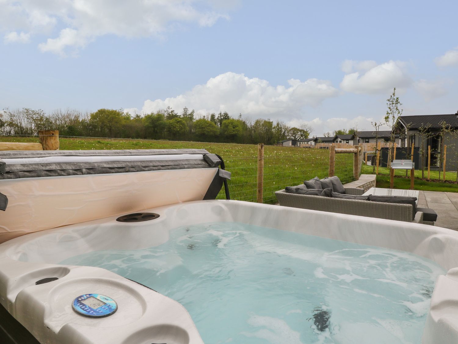 An outdoor hot tub with water and bubbles next to a patio with a sofa set in a fenced grassy area with buildings in the background at Lily in Broadwoodwidger near Ashwater