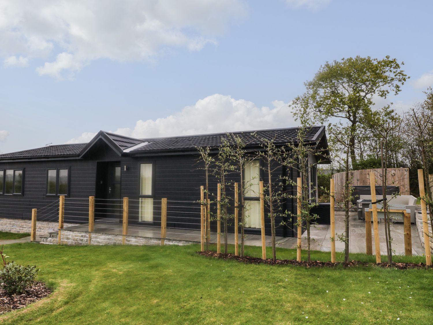 A detached single-storey black house with a tiled roof behind a wooden post fence on a grassy area with young trees at Lily in Broadwoodwidger near Ashwater