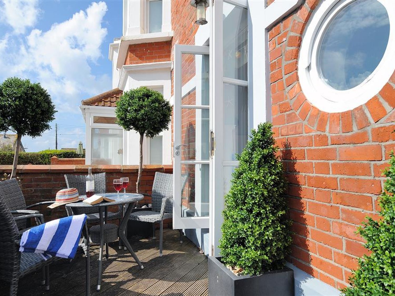 An outdoor patio with wicker chairs a table with two glasses of red wine a book and a hat near a brick wall with a circular window at Seaside Lyme Regis