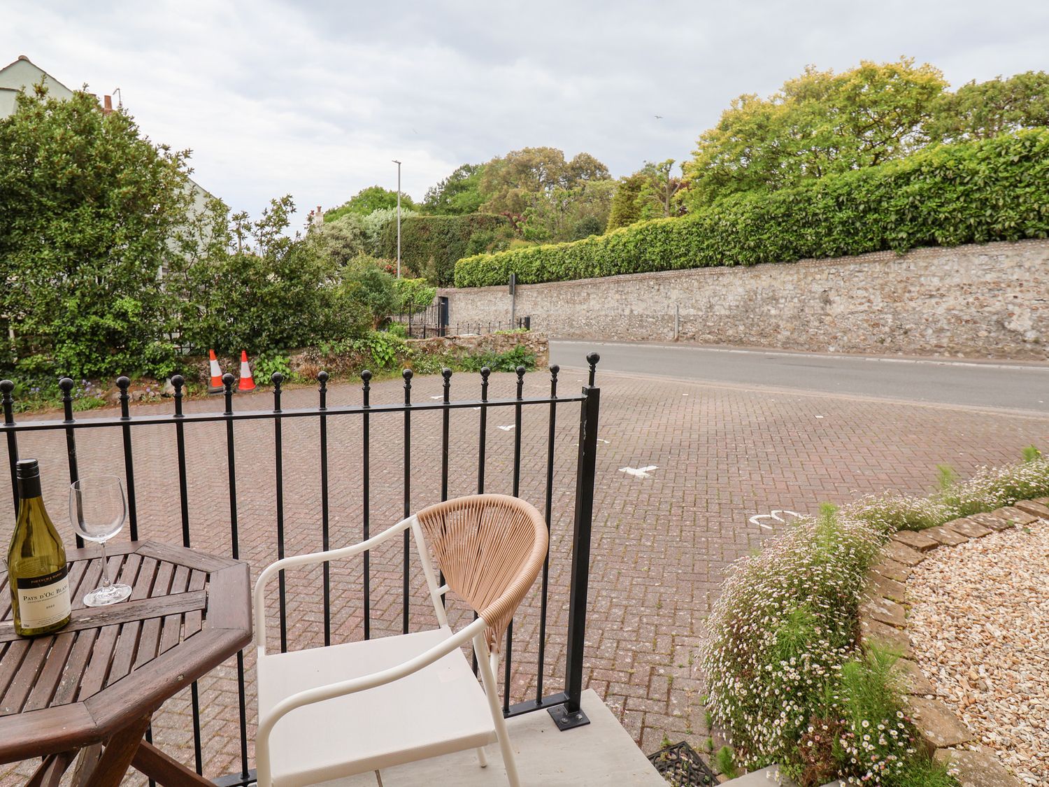 A small outdoor seating area with a table bottle of wine and glass near a road with hedges and stone walls at Sea Glass in Lyme Regis