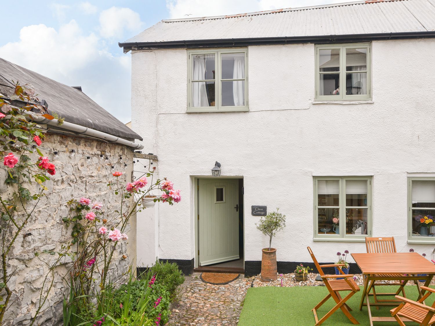 An outdoor patio area with wooden table and chairs next to a white cottage with green window frames and a green door with flowers along a stone wall at Cleve Cottage in Lyme Regis