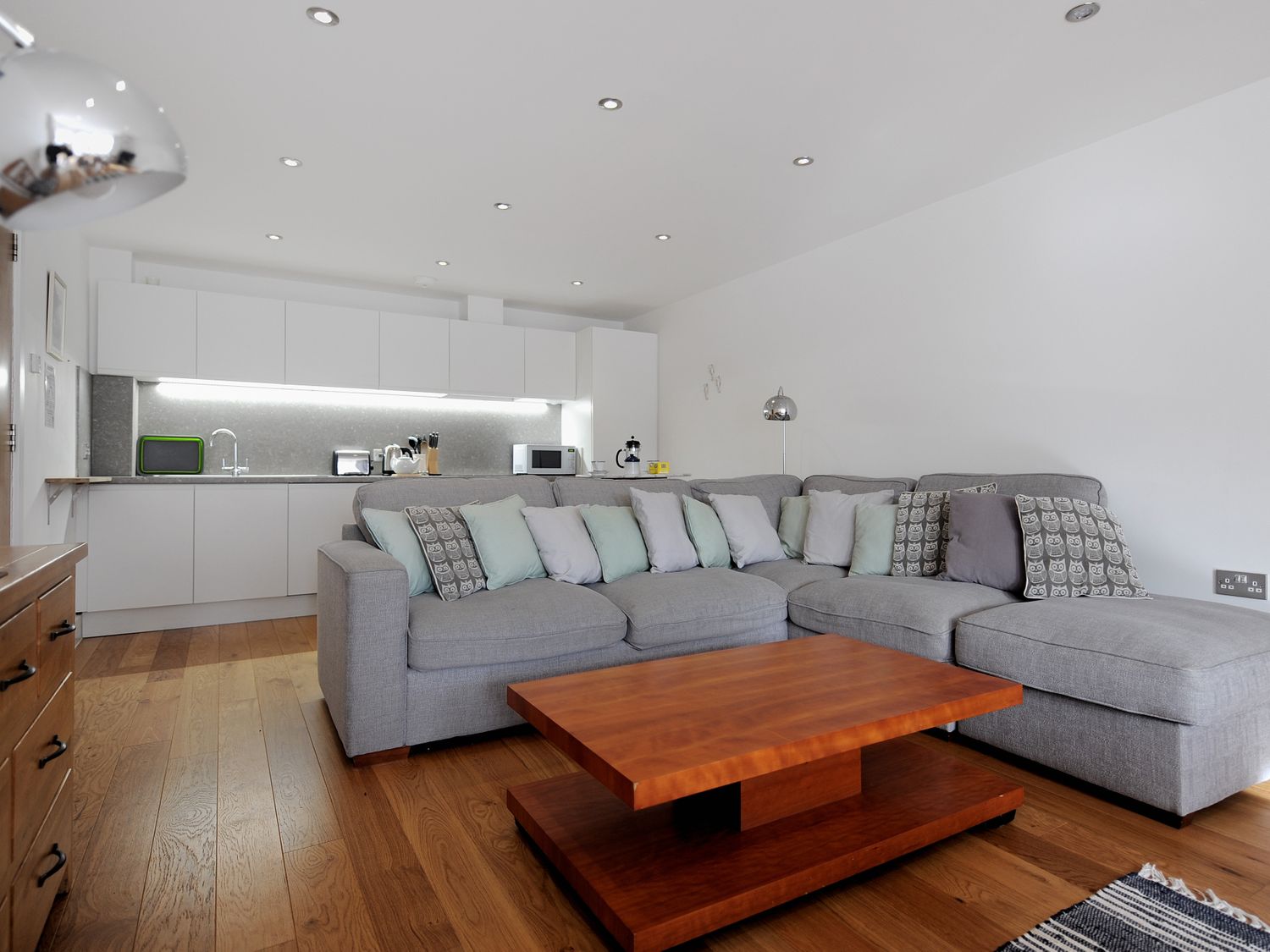 A living room with a grey sectional sofa and wooden coffee table next to a kitchen with white cabinets and appliances at Seashell in Lyme Regis