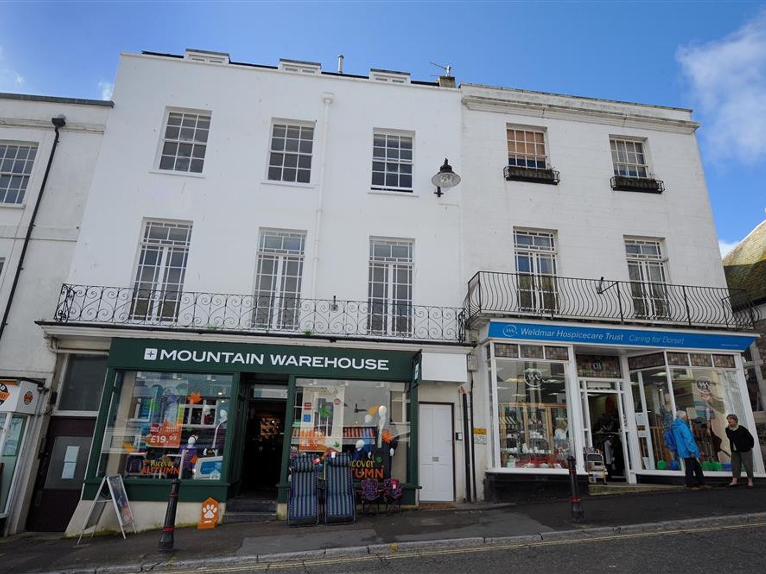 A street view of two white buildings with shops named Mountain Warehouse and Weldmar Hospicecare Trust with balconies above at Rooftops in Lyme Regis
