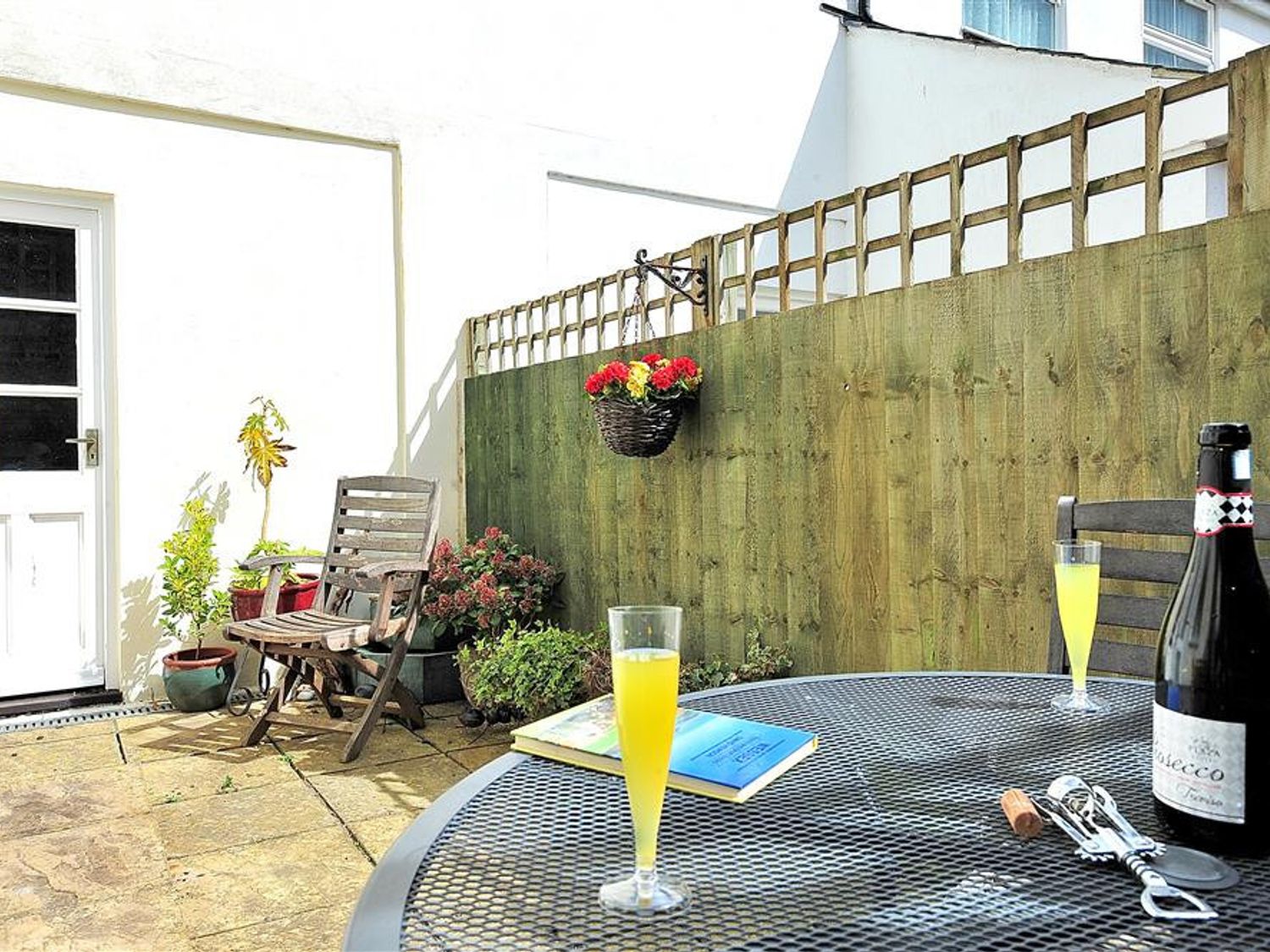 A patio area with wooden chairs potted plants a bottle of prosecco two glasses and a book on a metal table at Vine Cottage in Charmouth