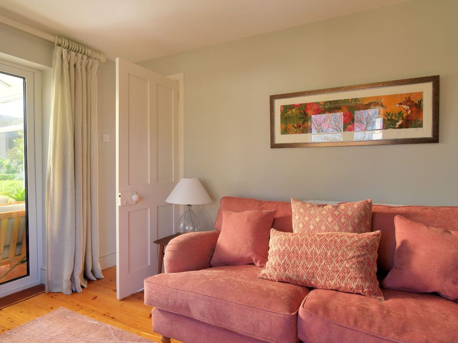 A living room with a pink sofa with cushions a table lamp an open door curtains and wall art at Hernelee Cottage in Lyme Regis