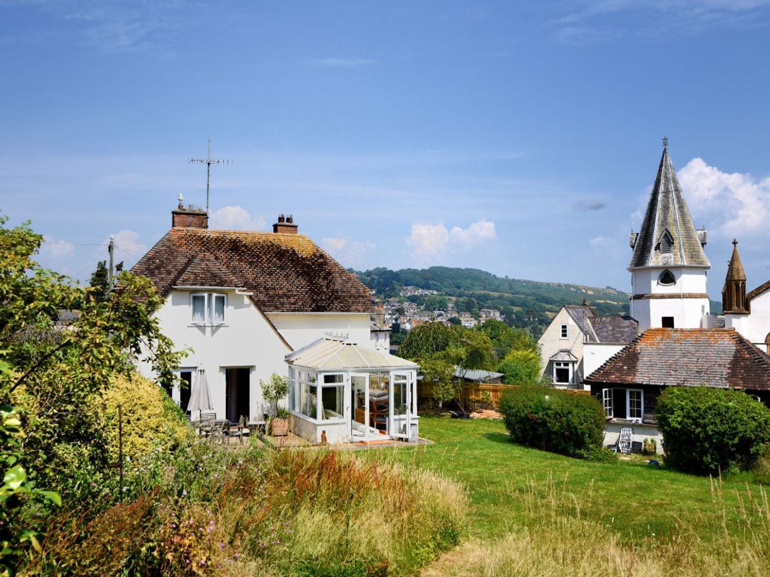 A house with a conservatory and a church tower in the background with grassy fields and bushes at Hernelee Cottage in Lyme Regis