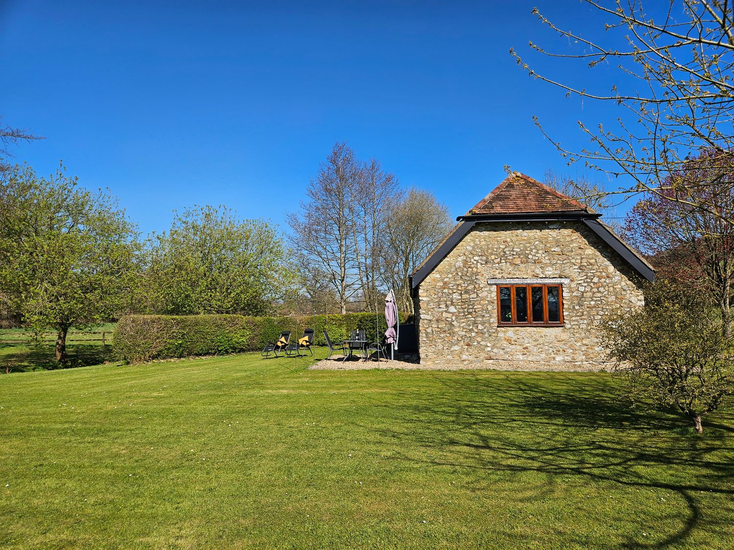 A stone cottage with a tiled roof and a window in a garden with lawn chairs and trees at The Cider House in Dalwood