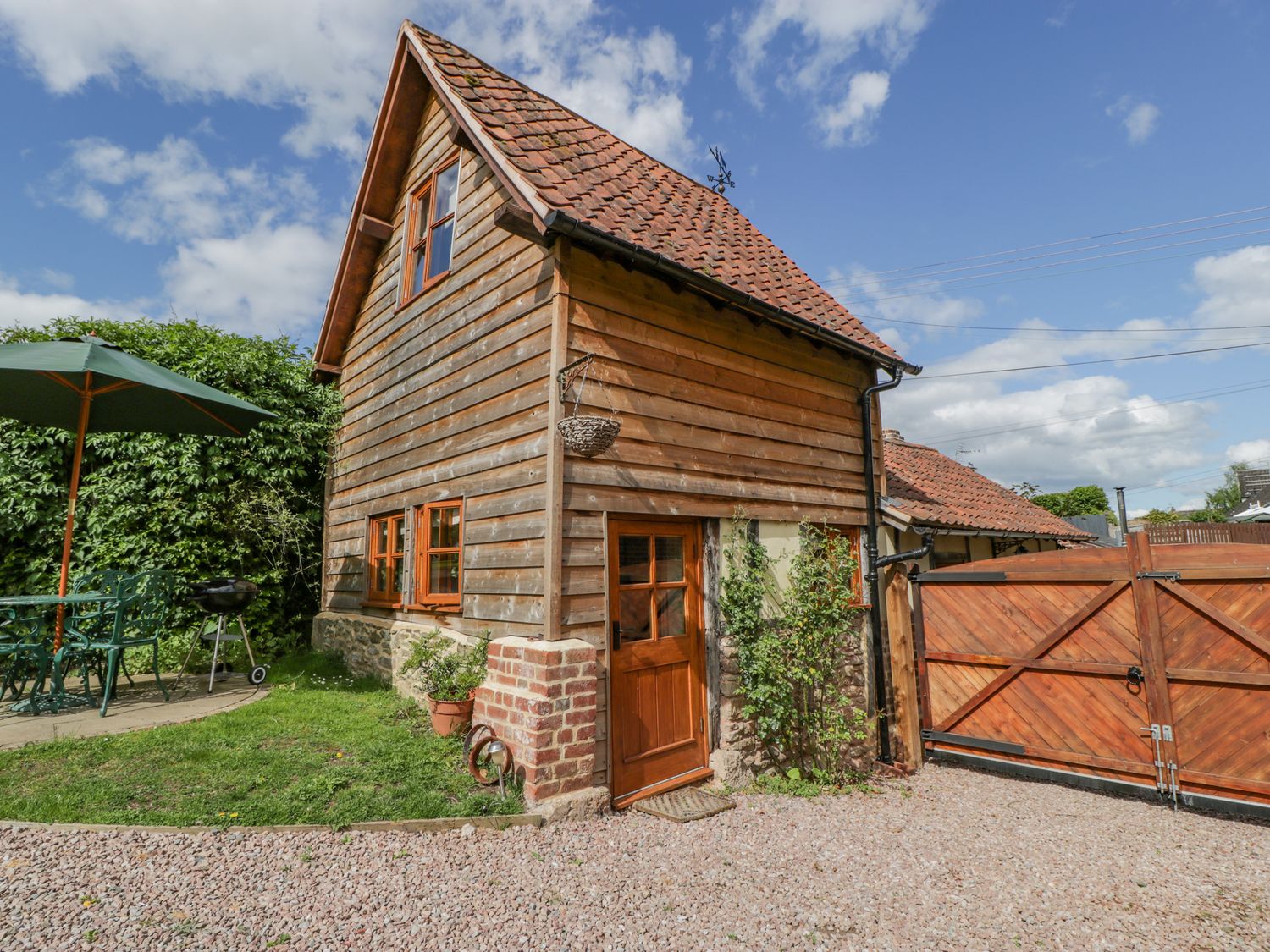 A wooden house with a tiled roof a gravel driveway a wooden gate and outdoor furniture with an umbrella at The Forge in Tarrington near Bartestree