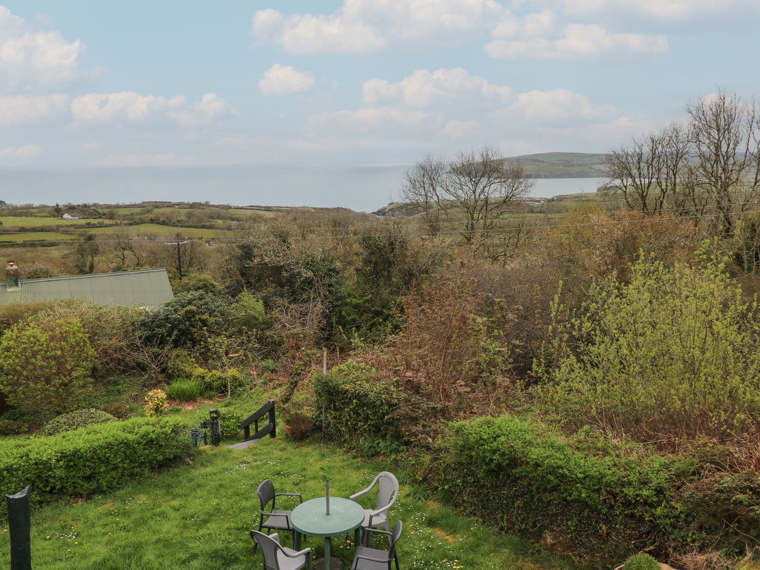 A garden with a round table and four chairs surrounded by bushes and trees with fields and hills in the background at Arfron in Dinas Cross
