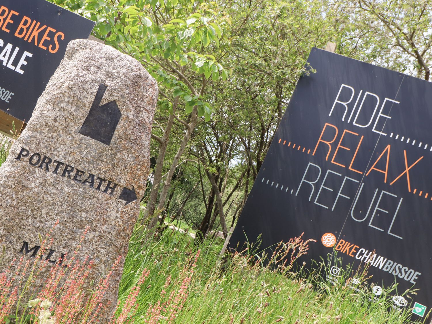 A stone signpost pointing to portreath and a black advertisement board for bike chain bissoe among grass and trees at 1 Woodlands Twelveheads near Chacewater