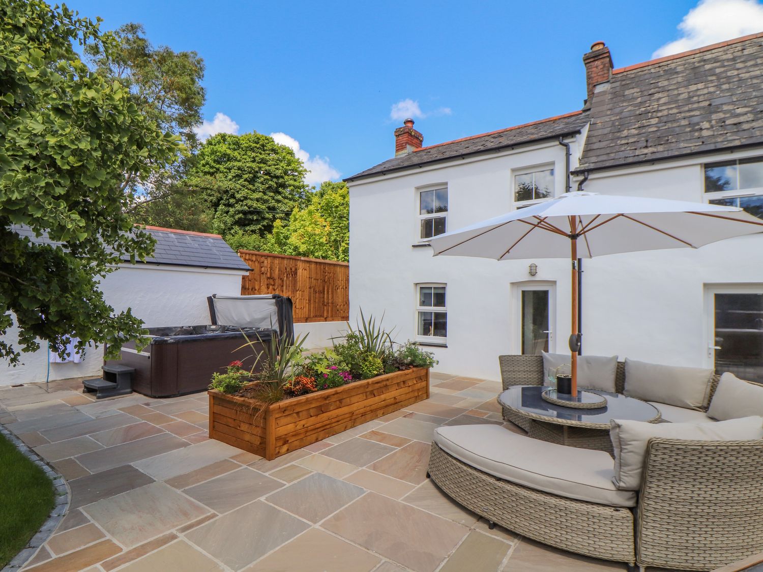 An outdoor patio area with a circular wicker sofa and umbrella a wooden planter and a hot tub at 1 Woodlands in Twelveheads near Chacewater