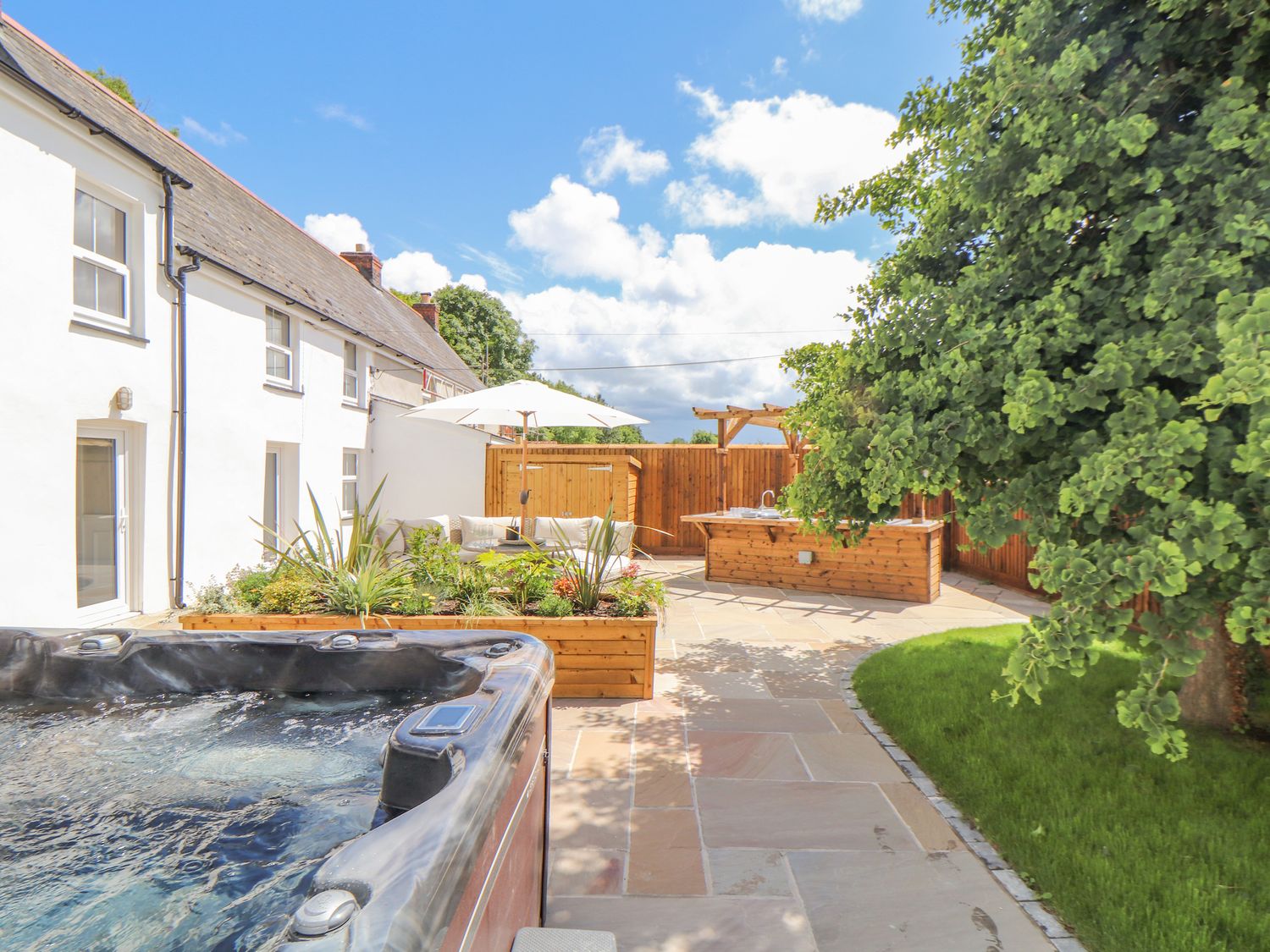 A backyard with a hot tub wooden planter with plants a seating area with an umbrella and a wooden outdoor kitchen at 1 Woodlands in Twelveheads near Chacewater