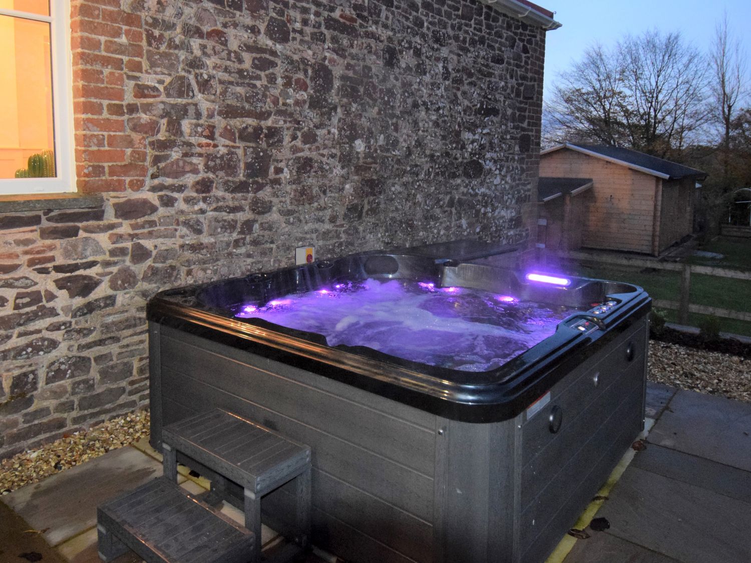 An outdoor hot tub with purple lights next to a stone wall and wooden shed at Hope House Stables in Witheridge