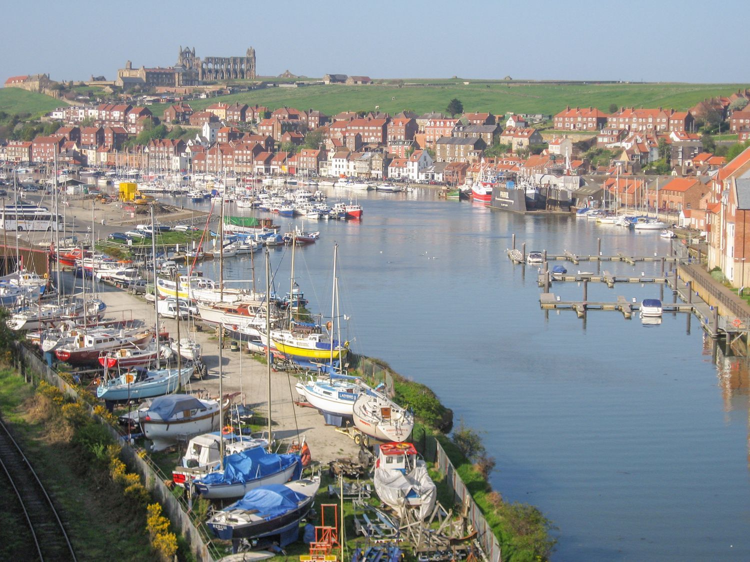 A harbor with boats docked and moored along the water with red brick buildings and a large historic abbey on a hill in the background at Fern House in Whitby