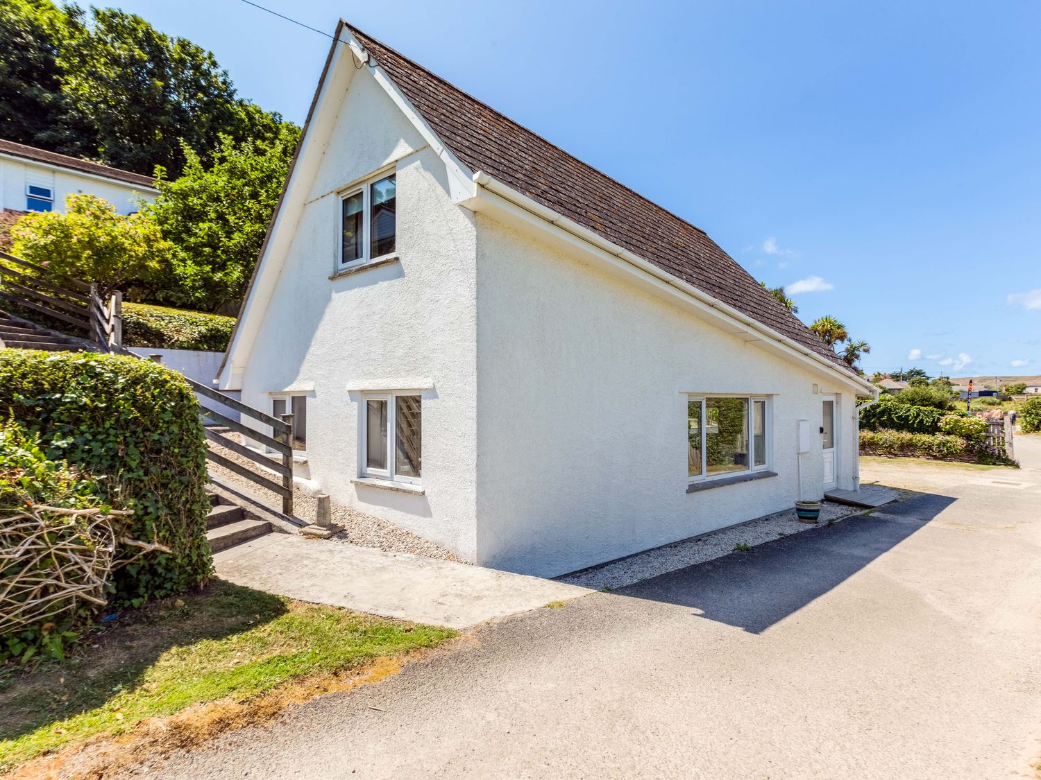 Exterior view of a white house with multiple windows and a sloping roof surrounded by bushes and trees at Baywatch Sands in Polzeath