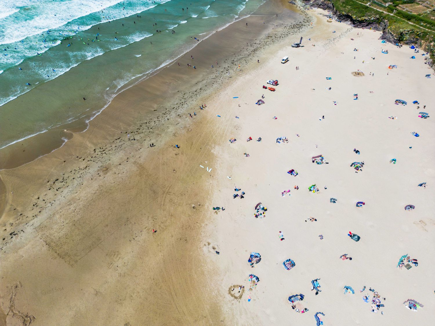 An aerial view of a beach with people on the sand and surfers in the water at Baywatch Sands in Polzeath