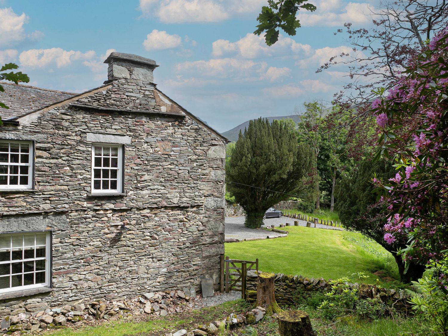 A stone house with windows near a grassy lawn and trees with purple flowers at Thorney How in Grasmere