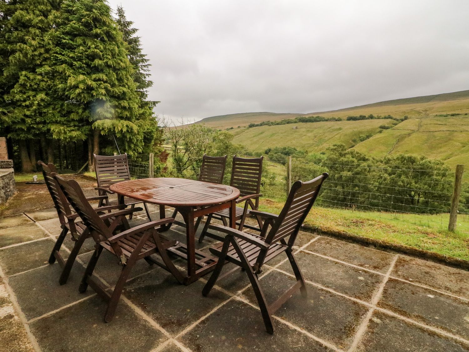 An outdoor patio with a wooden table and six chairs overlooking hills and trees at Garth Gill in Garsdale near Hawes