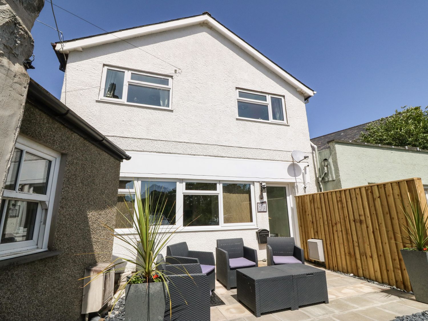 A patio with black wicker chairs and tables next to a white two story house with windows at Gwel Afon in Llanystumdwy near Criccieth