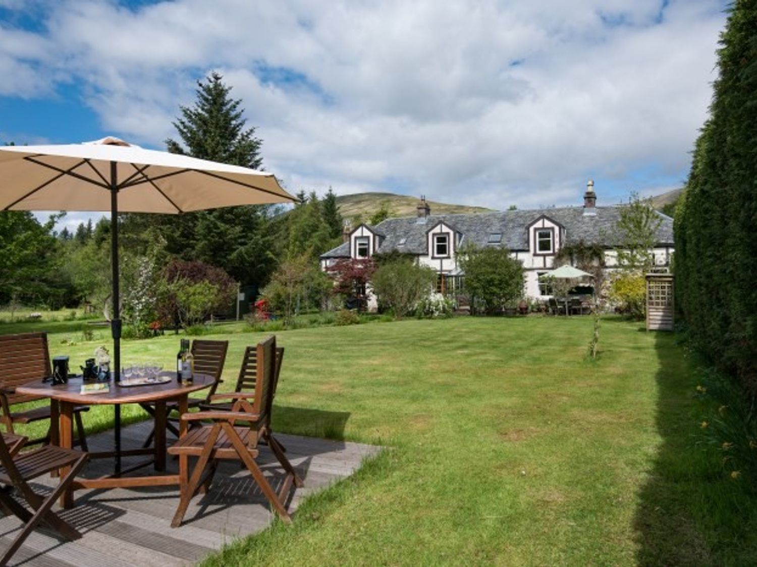 A backyard with wooden patio furniture and an umbrella in front of a house at Strone House in Loch Lomand & Trosschs National Park
