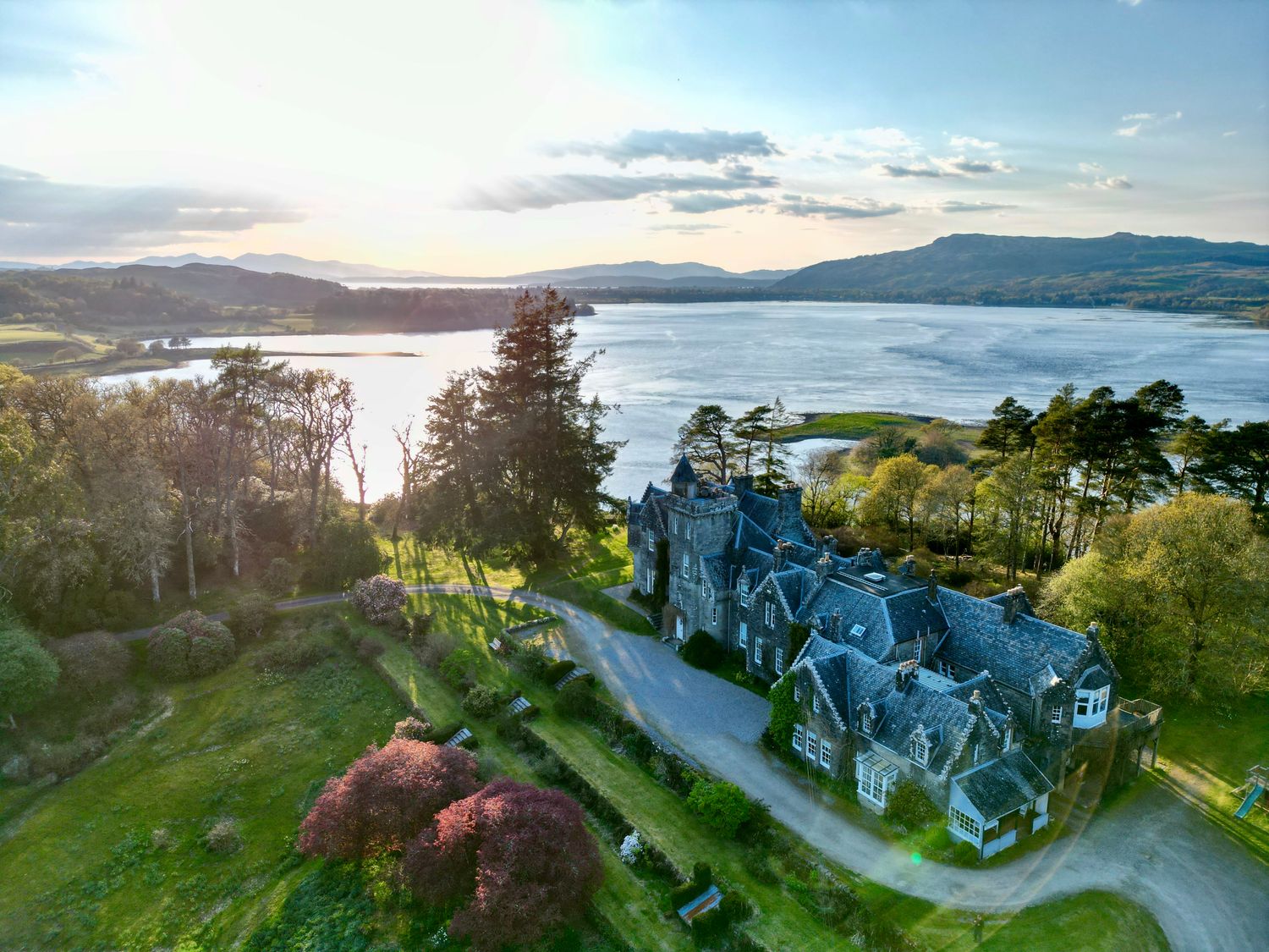 An aerial view of a large stone house with multiple roofs near a body of water surrounded by trees and hills at Achnacloich in Connel
