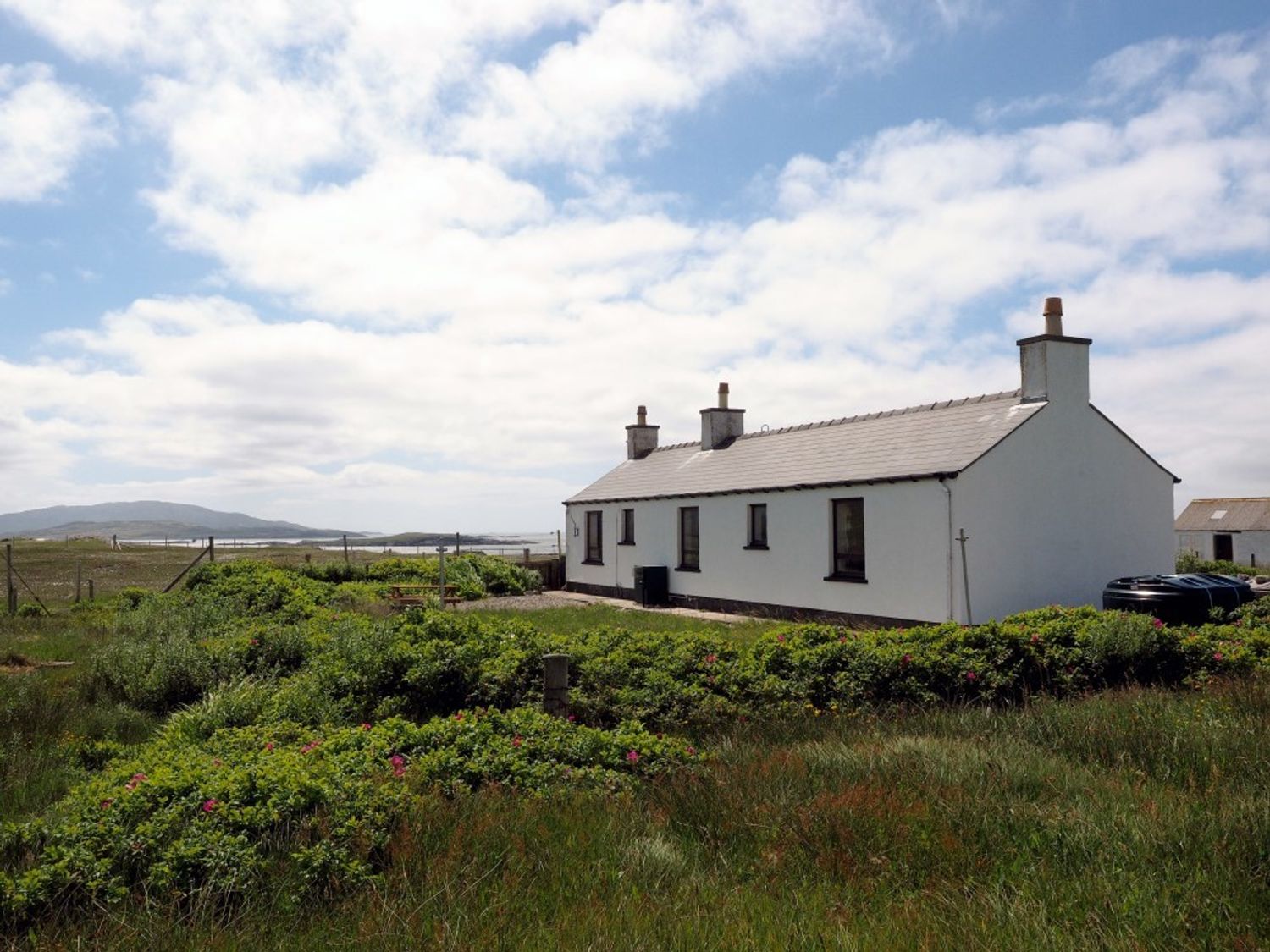 A white cottage with chimneys surrounded by grass and bushes under a partly cloudy sky at Spindrift Breanais nr Timsgearraidh