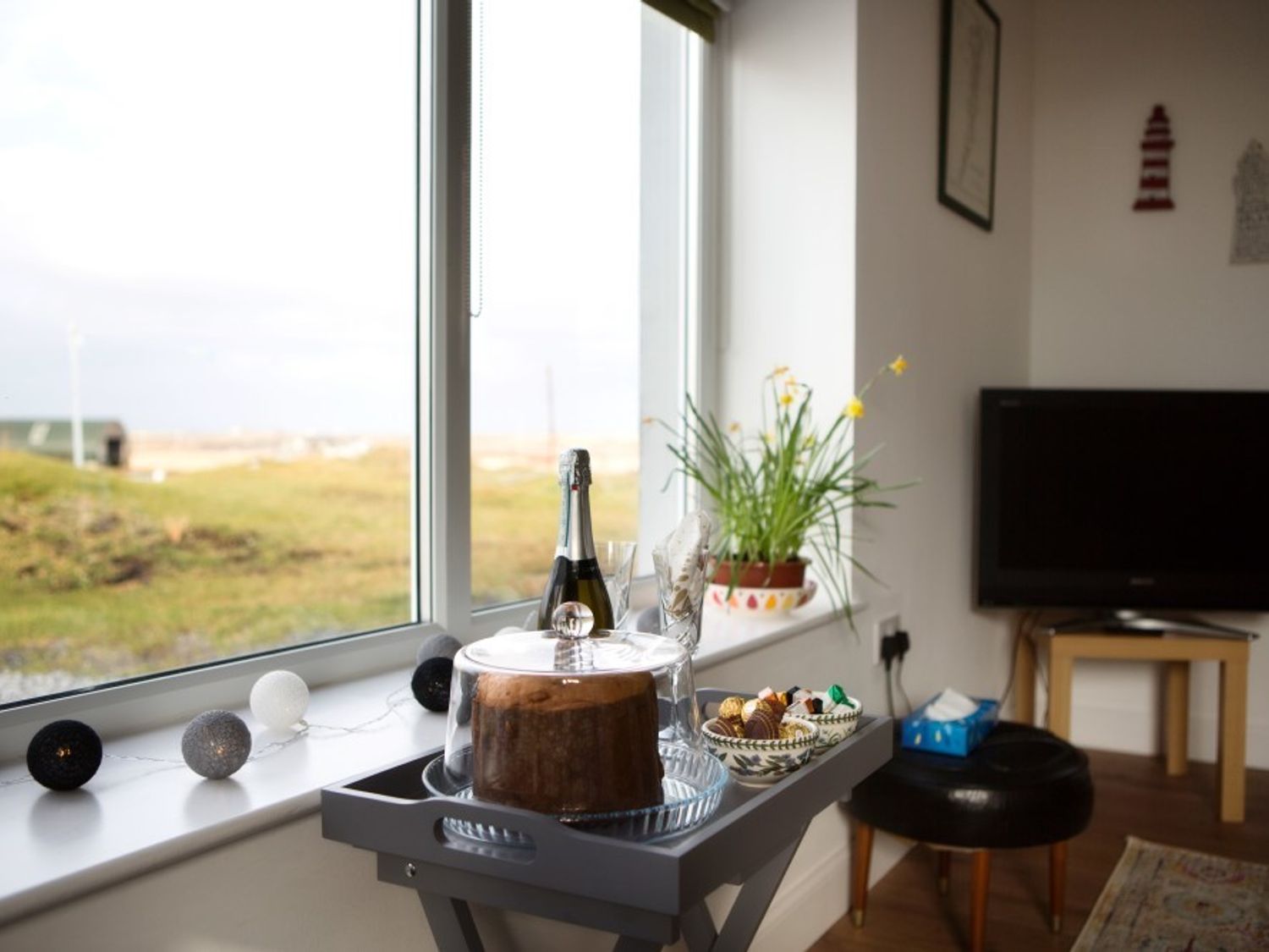 A window with a tray holding a cake, champagne bottle, glasses, and snacks in a room with a television and plant at Ceannard Cottage in Daliburgh