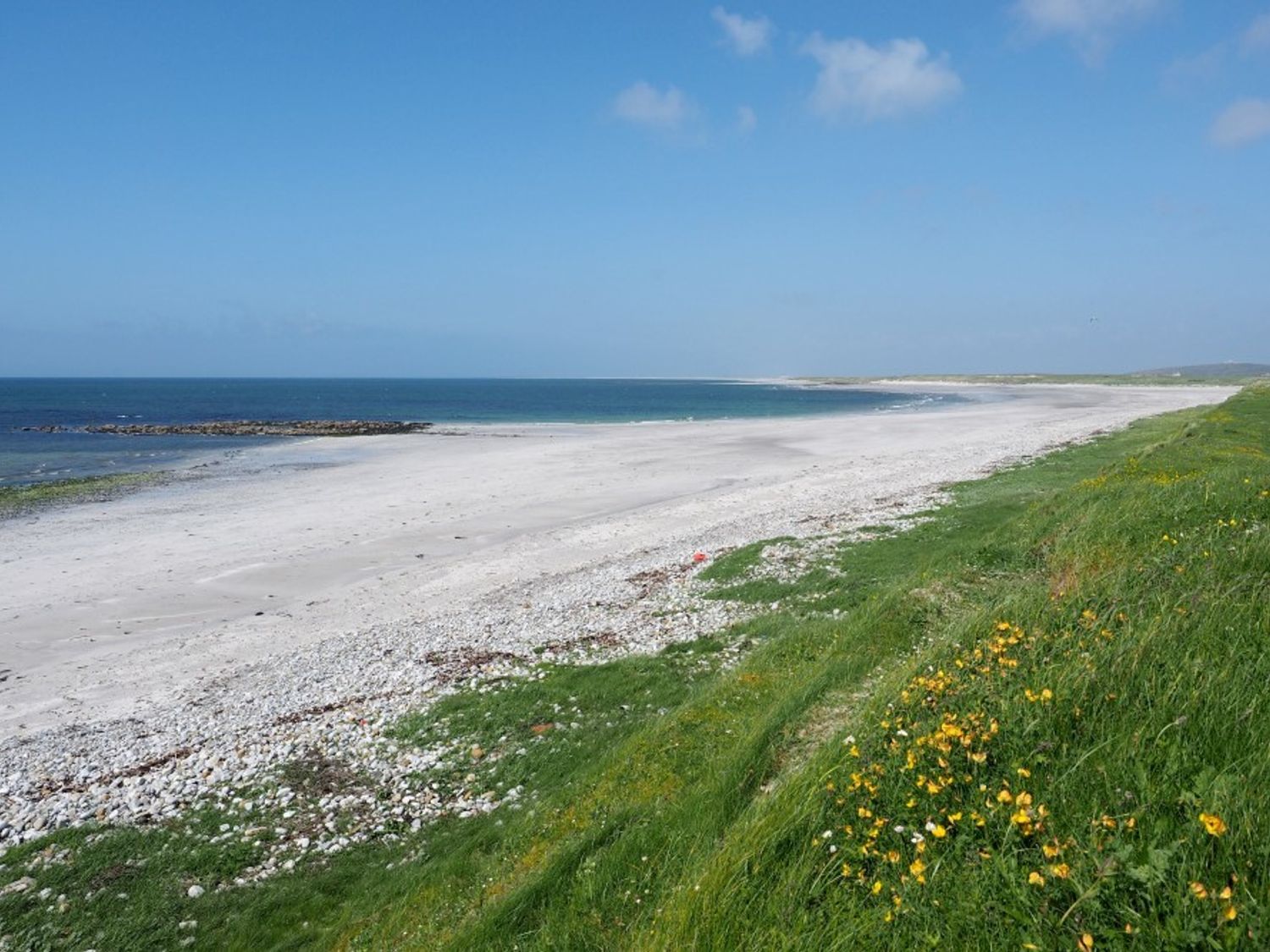 A sandy beach with pebbles and green grass with yellow flowers by the sea at Taigh Dhonnchaidh Peninerine near Creagorry
