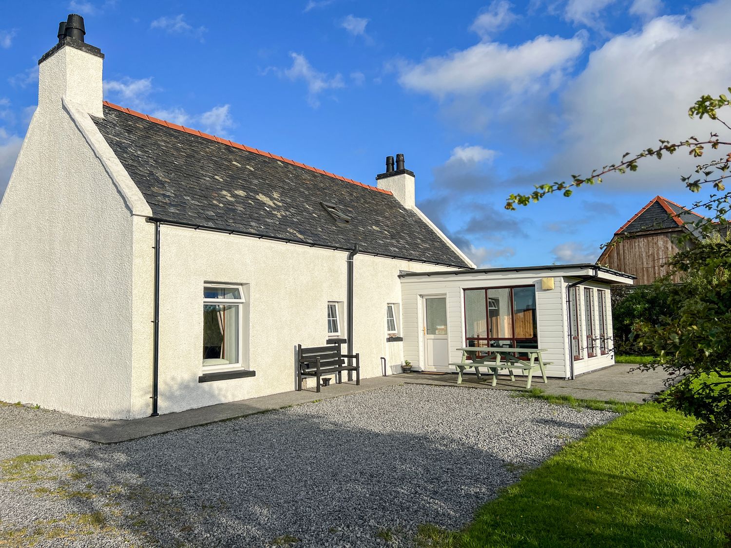 An exterior view of a white cottage with a slate roof a gravel driveway a picnic table and a bench at Corncrake Cottage SU in Daliburgh