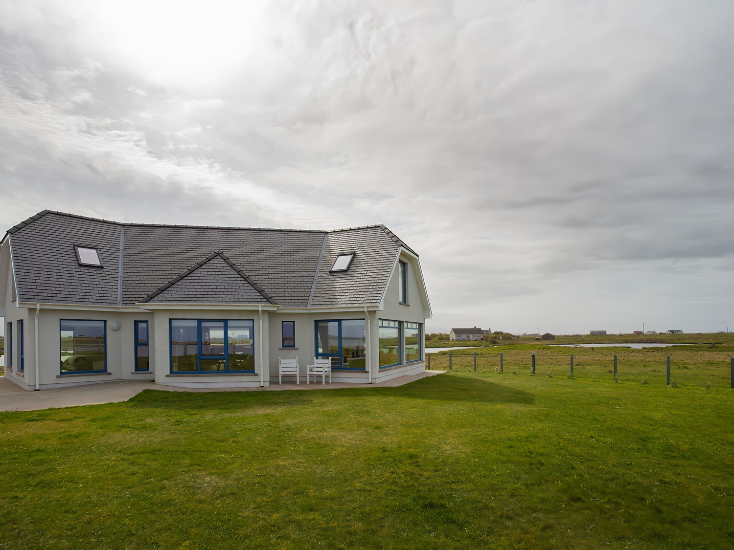 A house with multiple windows and two white chairs on the patio surrounded by a grassy field at Gualan Sands in Iochdar