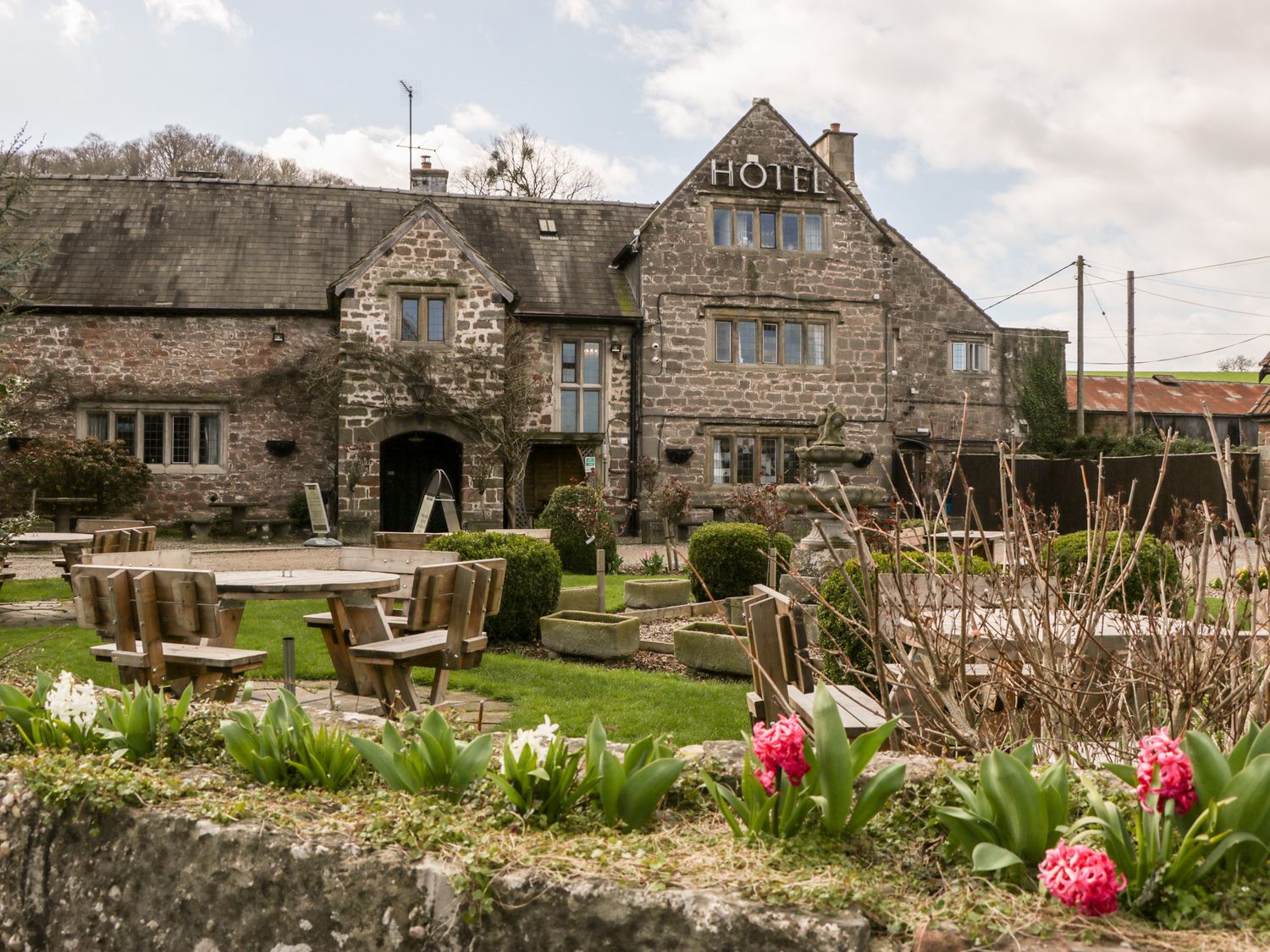 An outdoor seating area with wooden tables and chairs in front of a stone hotel building with flowers and shrubs in the garden at Steel Works Farm in Gorsley
