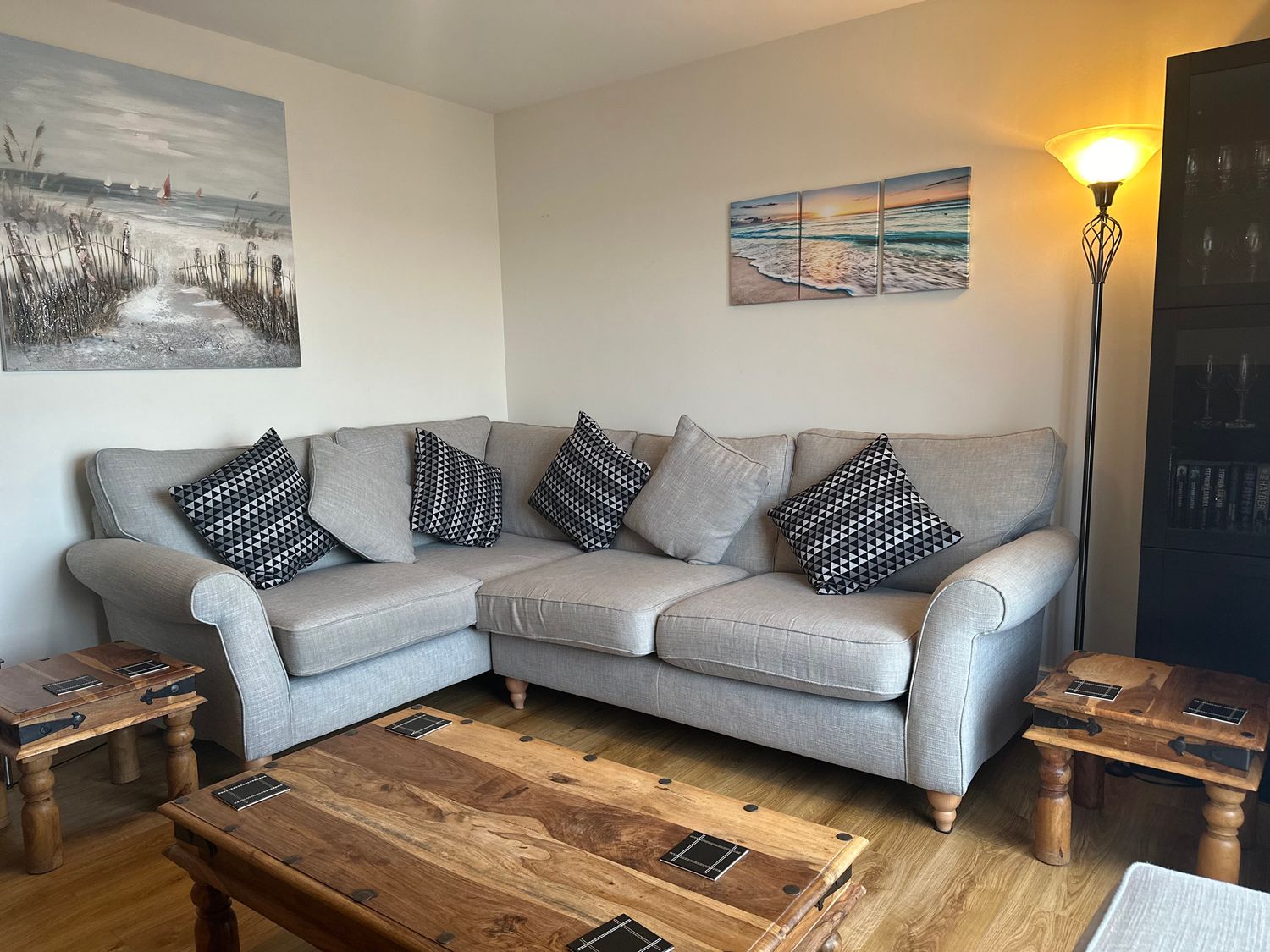 A living room with a grey corner sofa with patterned cushions wooden tables a floor lamp and beach-themed wall art at Apartment 3 in Trearddur Bay