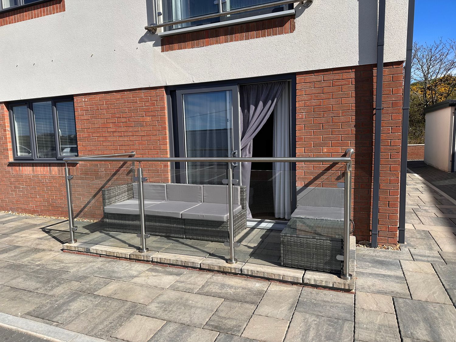 An outdoor patio area with a cushioned sofa and glass railing outside a brick building at Apartment 3 in Trearddur Bay